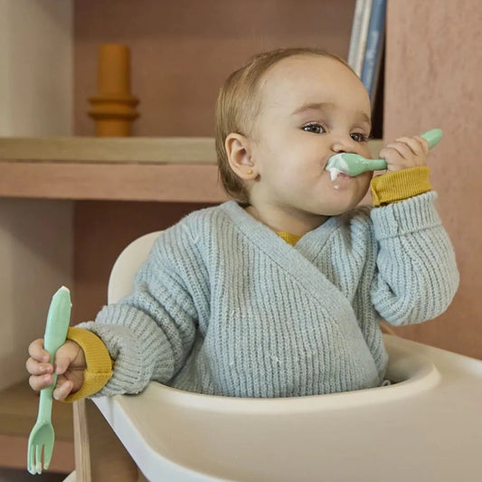 Baby sitting in a high chair with green utensils, wearing a light blue knitted sweater. — BambiniJO Jordan