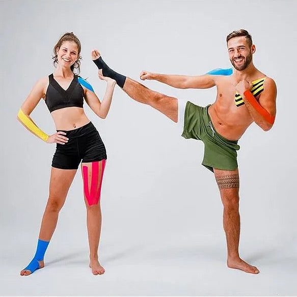 A man and a woman demonstrate athletic compression sleeves and leg wraps in a studio setting. The man wears green athletic shorts and extends his leg while wearing blue, yellow, and black compression sleeves. — BambiniJO Jordan